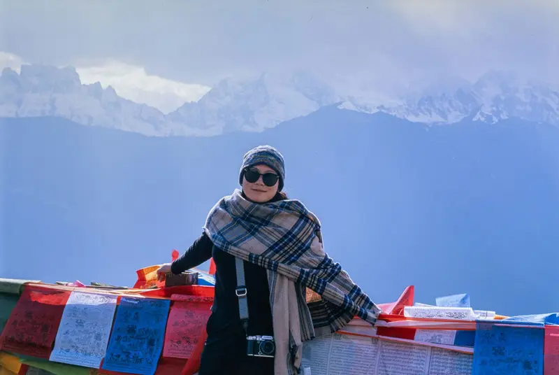 Woman in sunglasses beside prayer flags, snow peaks behind